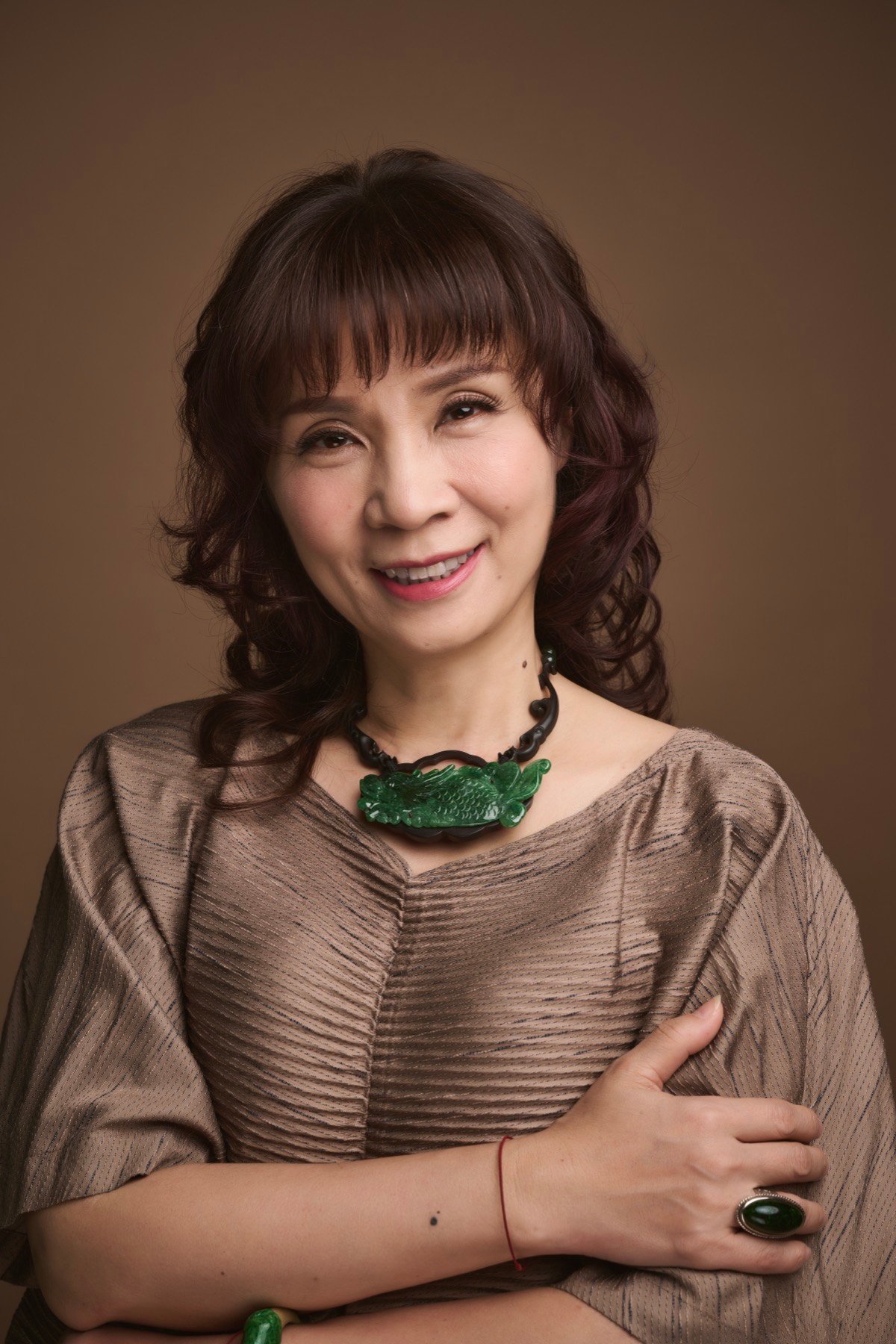 王牧絃 Jenny，身著金棕色壓褶上衣與翡翠珠串項鍊，環抱雙臂微笑